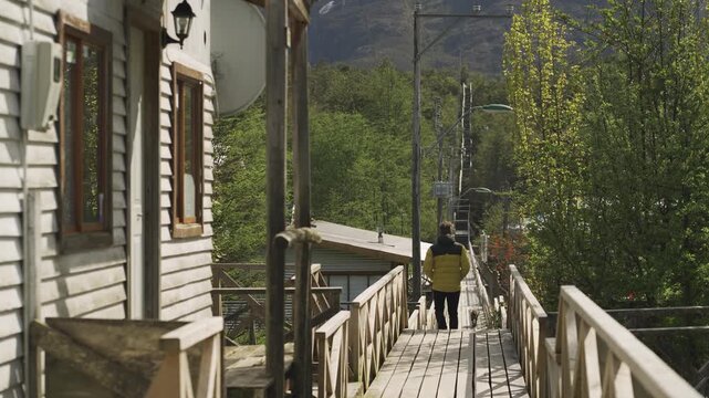 Person in a yellow jacket walks along a wooden boardwalk beside a white house, surrounded by trees and mountains in a rural setting.