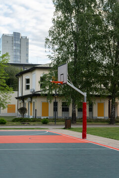 Empty outdoor basketball court features a backboard with an orange hoop on a steel pole with red safety padding against a public building green trees and highrise skyscraper background area.