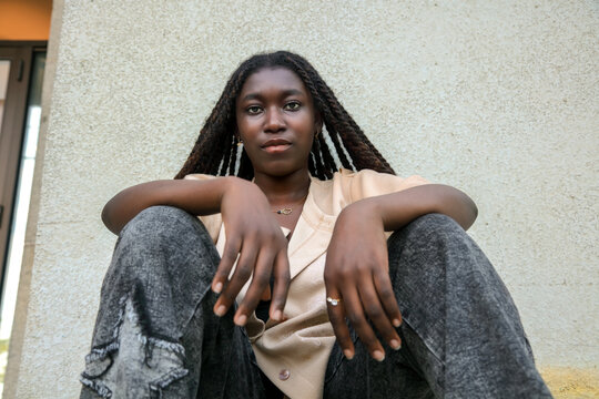 Urban portrait of a cool young Black woman sitting with a relaxed attitude against a concrete wall