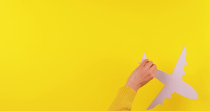 Close-up video of a hand holding a white airplane symbol moving in and out of the frame on a yellow background. Travel and aviation concept symbolizing flights, journeys, tourism, and air transport.