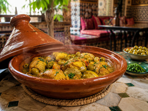  Taj&iacute;n tradicional con patatas doradas, aceitunas verdes, verduras y abundantes hierbas arom&aacute;ticas, presentado en una cazuela de barro dentro de un acogedor restaurante de estilo marroqu&iacute;.