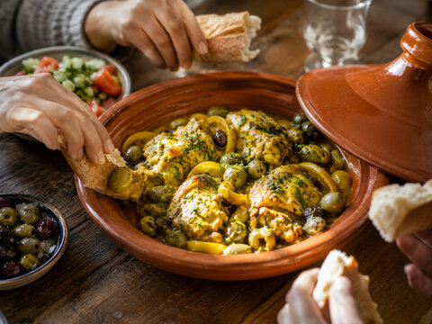 Taj&iacute;n tradicional con abundantes aceitunas verdes, hierbas arom&aacute;ticas y salsa especiada, servido en una cazuela de barro mientras varias personas comparten pan alrededor de la mesa.