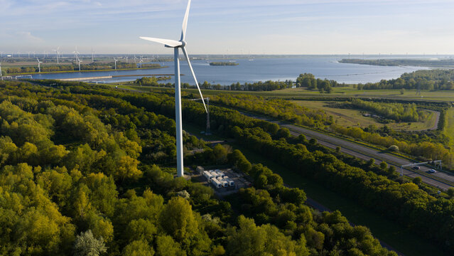 Aerial view of a wind turbine standing amidst lush green trees near a highway and the coastal waters in Bruinisse, Zeeland, Netherlands.