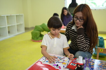 Naklejka premium An Asian kindergarten teacher with glasses patiently guides a young boy during an interactive learning activity at a desk in a classroom, highlighting early childhood mentorship and education.