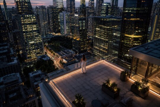 High angle view of a woman standing on a luxury high-rise rooftop terrace overlooking a glowing night city skyline. Concept of success, leadership, and premium real estate.