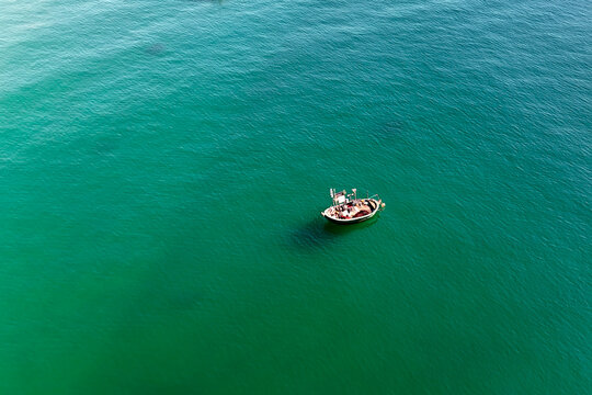 Small fishing vessel rests on calm, deep green ocean water. Sunlight reflects off the surface, casting a clear shadow below