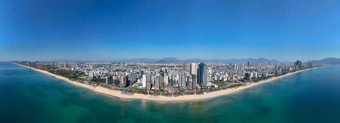 Fototapeta premium Expansive aerial view of Da Nang city buildings lining the vast My Khe Beach with turquoise ocean under a bright blue sky. Mountains rise in the distance