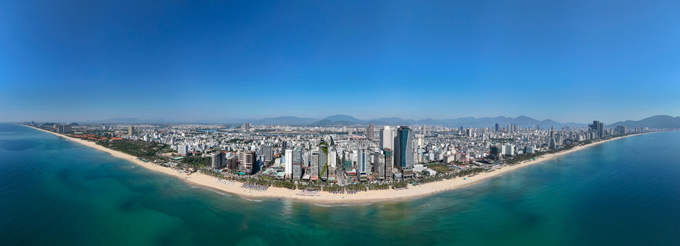 Expansive aerial view of Da Nang city buildings lining the vast My Khe Beach with turquoise ocean under a bright blue sky. Mountains rise in the distance