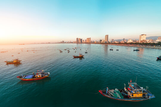 Fishing boats float on calm, deep blue ocean water offshore from a busy city beach lined with tall buildings at sunset