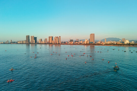 Numerous paddleboarders and kayakers enjoy water activities on the calm bay off the Da Nang cityscape, with distant boats visible in the morning light
