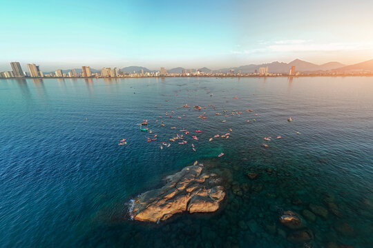 A large group of stand up paddleboarders and boats enjoy the clear ocean waters around a prominent rock