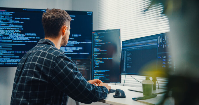 Young Caucasian male programmer writes code using multiple screens in home office workspace. AI development, cybersecurity, network database system, software engineering, remote tech startup concept.
