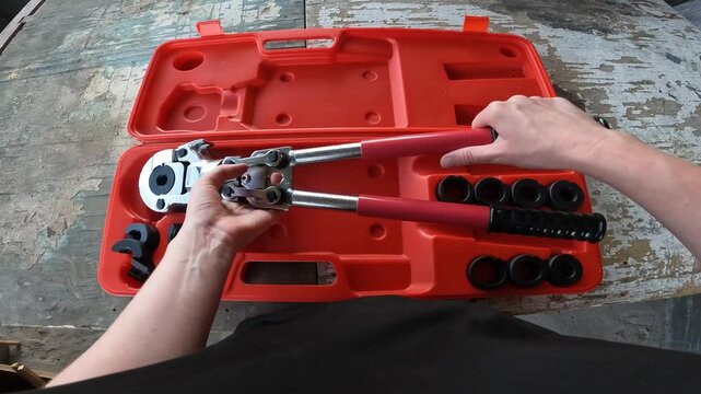 Manual crimping pliers for metal-plastic pipes in a case. Plumber holds a professional crimping tool for installing PEX pipes. Top view of a case with interchangeable nozzles on a wooden background