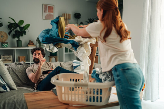 Irritated woman throwing laundry at lazy man avoiding housework on sofa, causing conflict and relationship struggle