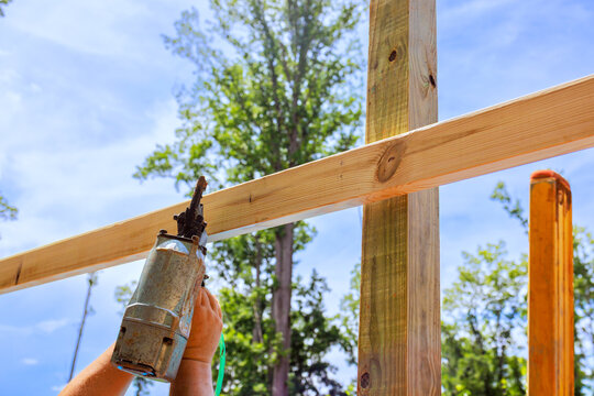 Worker fastens wooden beams with pneumatic nail gun while constructing structure with trees nearby.