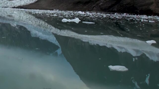 An immense Arctic glacier stands watch. Its icy face shimmers over tranquil, deep emerald water, dotted with pristine floating ice fragments. A serene, powerful spectacle unfolding in the far north.