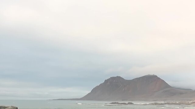 Witness the raw power of Arctic Svalbard, Norway, as mighty waves crash against ancient, rugged shores. Overcast skies loom above, creating a dramatic atmosphere. Majestic mountains rise from the sea.