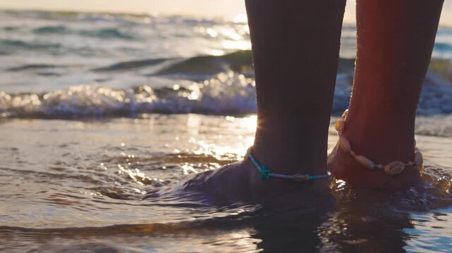 A young woman stands motionless in the shallows. gentle waves lap at the shore, leaving behind fine spray, ripples, and trickles of water around her colorful bracelets of beads and shells.