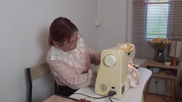Elderly woman with measuring tape around neck wearing glasses uses sewing machine clothes at home. Senior tailor works creative hobby during retirement lifestyle.