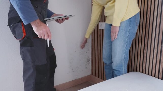 Worried homeowner showing an expert mold and fungus on bedroom wall caused by dampness, while professional inspector takes notes on tablet to prepare quote.