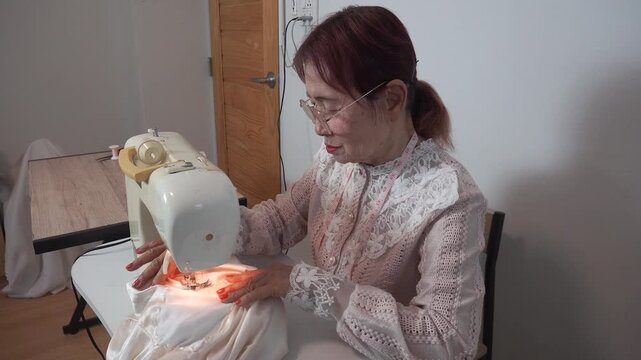 Elderly woman with measuring tape around neck wearing glasses uses sewing machine clothes at home. Senior tailor works creative hobby during retirement lifestyle.