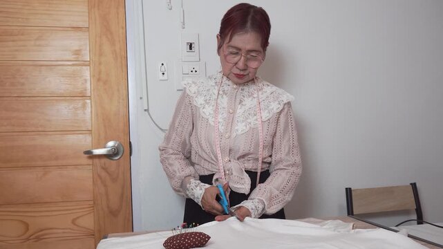 Elderly woman with measuring tape around neck uses scissors to cut clothes at home. Senior tailor works her creative hobby during retirement lifestyle.