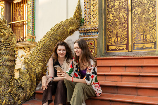Two Women Sit on Steps and Check Their Phones at a Temple