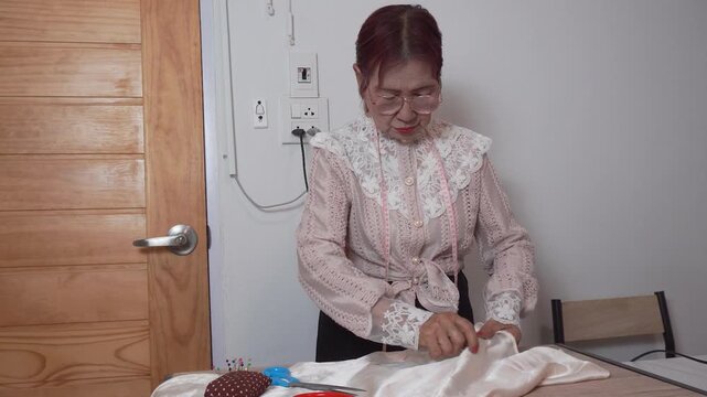 elderly woman with measuring tape around neck wearing glasses uses needle for sewing clothes at home. Senior tailor works her creative hobby during happy retirement lifestyle