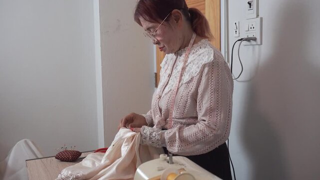 elderly woman with measuring tape around neck wearing glasses uses needle for sewing clothes at home. Senior tailor works her creative hobby during happy retirement lifestyle