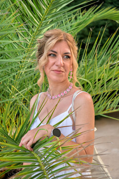 ​Attractive caucasian woman in white dress posing behind green palm leaves, summer vacation lifestyle