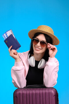 Smiling young woman wearing sunglasses holding passport and boarding pass with suitcase ready for vacation
