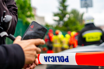 A witness or reporter recording a car accident scene with a smartphone from behind a red and white police cordon tape with the text "POLICJA", with a heavily blurred background of firefighters and eme © Adam