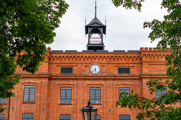 Facade of a historic, revitalized factory building in Łódź, built from red brick, featuring a central analog clock, an ornamental metal turret on the roof, and black multi-paned windows, framed by gre © Adam