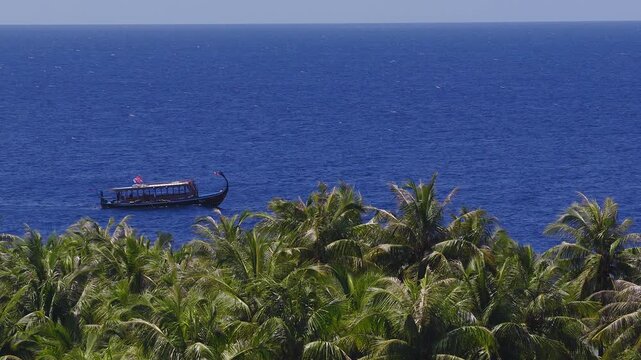 Aerial wide view over coconut palms to the Indian Ocean at Dhigurah, Maldives. A curved prow dhoni drifts near shore under bright midday sun with small flags fluttering.