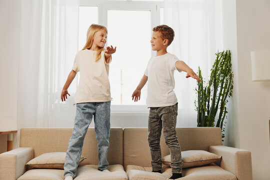 Two happy children jumping on a beige sofa in a bright living room, enjoying playful moments and sibling fun together at home.
