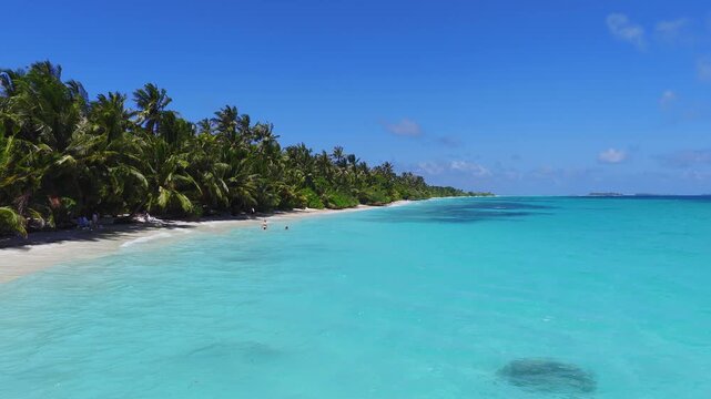 Aerial view of Dhigurah Island, Maldives, with palm lined white sand meeting a turquoise lagoon, dark reef patches, a round coral bommie, and people as the camera glides