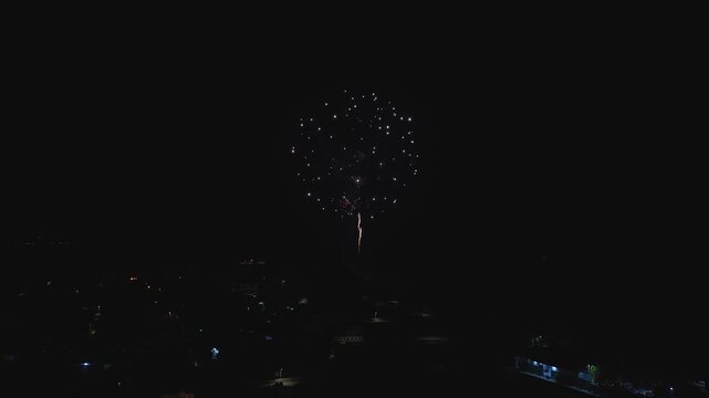 Aerial drone view shows night fireworks over Dhigurah Island, Maldives. Bright trails rise and bursts bloom as streetlamps and house lights map the shoreline below.