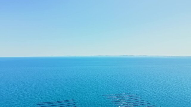 Mussel farm is located in the ocean with rows of long lines set in the water. Bright sky shows few clouds as sunlight reflects off the surface. Distant land can be seen.