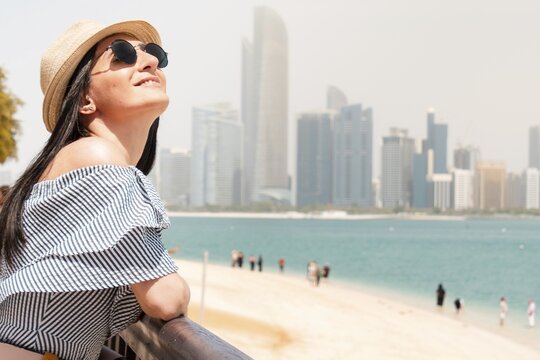Happy young female tourist on viewpoint in hot day visiting Abu dhabi, UAE - Traveler girl taking portrait in front of capital skyline background. Urban middle east city life travel attractions