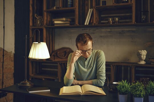 In a serene study space, a thoughtful man sits at a desk, deeply engaged in reading a book. Soft light from a lamp creates a warm ambiance, highlighting wooden shelves filled with decor