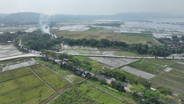 Aerial view of the rural landscape featuring rice fields, a road with traffic, and smoke in Ambarawa, Central Java, Indonesia.