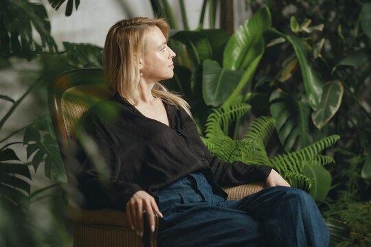 A woman is seated comfortably in a stylish chair amidst a vibrant collection of indoor plants. She gazes thoughtfully outside, enjoying the peaceful ambiance of her surroundings
