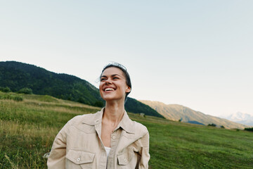 Fototapeta premium Smiling woman in casual jacket standing outdoors in a green field with mountains in the background. Concept of lifestyle, nature, happiness, and freedom in soft natural light.
