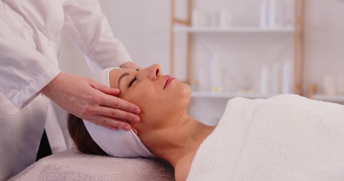Side view of young woman client having face massage, receiving beauty procedure from female beautician in beauty clinic. Cosmetologist performing rejuvenating beauty skincare treatment in spa salon.