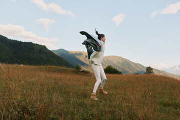Fototapeta premium Young woman jumping in a grassy field with mountains in the background. Outdoor lifestyle concept with casual clothing and natural light during daytime.