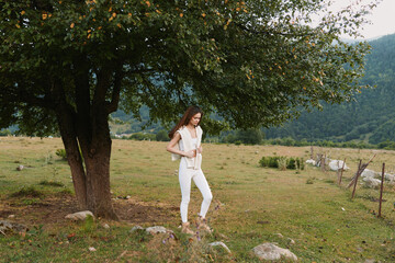 Fototapeta premium Woman standing beside a large tree in a rural meadow, wearing white clothing. Lifestyle portrait outdoors, peaceful countryside scene, natural light, relaxed pose, simple fashion.