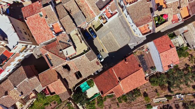 Rooftops and streets of a small town are shown from above in sunny weather. Various buildings are visible along with patches of green space and parked cars.