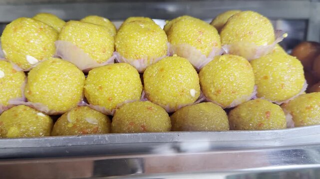 Close-up 4K video of bright orange Indian laddus arranged in a glass display at a sweet shop, showcasing their glossy texture, traditional dessert appeal, and festive food presentation.