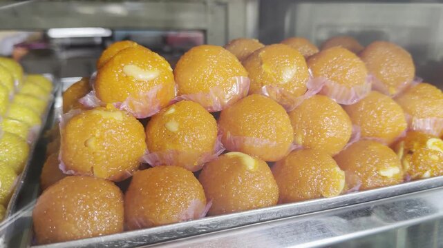 Close-up 4K video of bright orange Indian laddus arranged in a glass display at a sweet shop, showcasing their glossy texture, traditional dessert appeal, and festive food presentation.