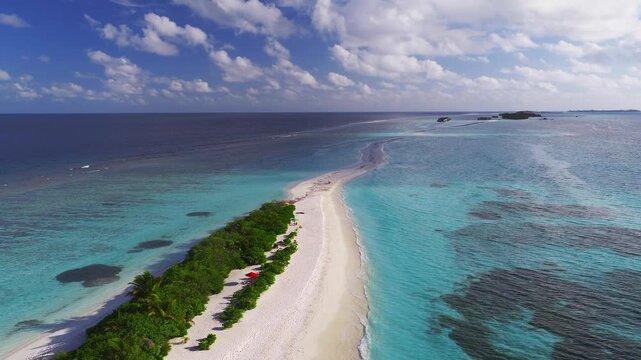 Aerial view shows Dhigurah Island, Maldives, long white sand spit curving into turquoise lagoon with coral patches, midday light, clouds, gentle waves, slow sweeping motion